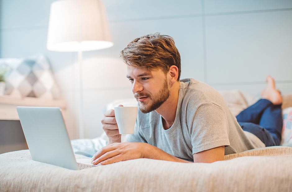 Man som ligger på sängen dricker en varm dryck medan han läser e-post på sin dator